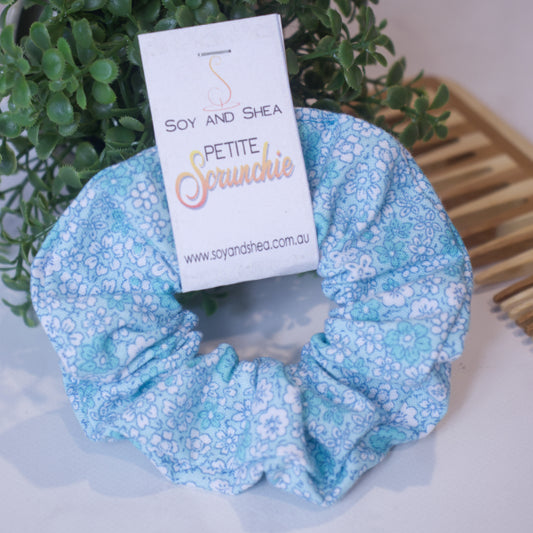 A colourful scrunchie with a cardboard tag rests against a green plant with a wooden comb in the background.  The Scrunchie is pale blue and covered in small white and teal forget-me-not flowers