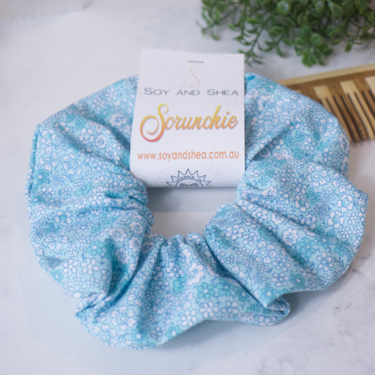 A colorful scrunchie sits on wooden surface resting against a wooden comb with greenery in the background.  A cardboard tag reads SOY AND SHEA SCRUNCHIE.    The scrunchie is pale blue with white and teal forget-me-not flowers tightly covering it.