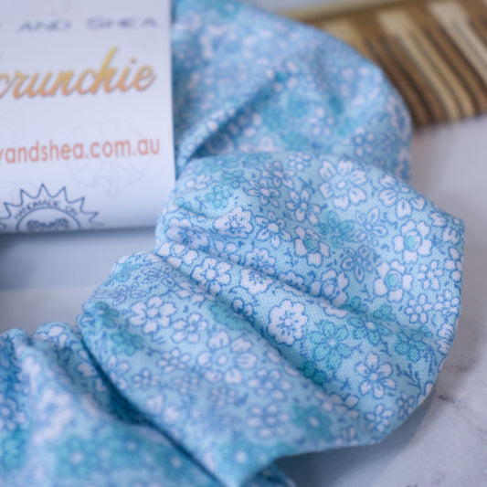 A close up of the fabric design on the Forget-me-not scrunchie showing the details of the the small white and teal flowers against a pale blue background.