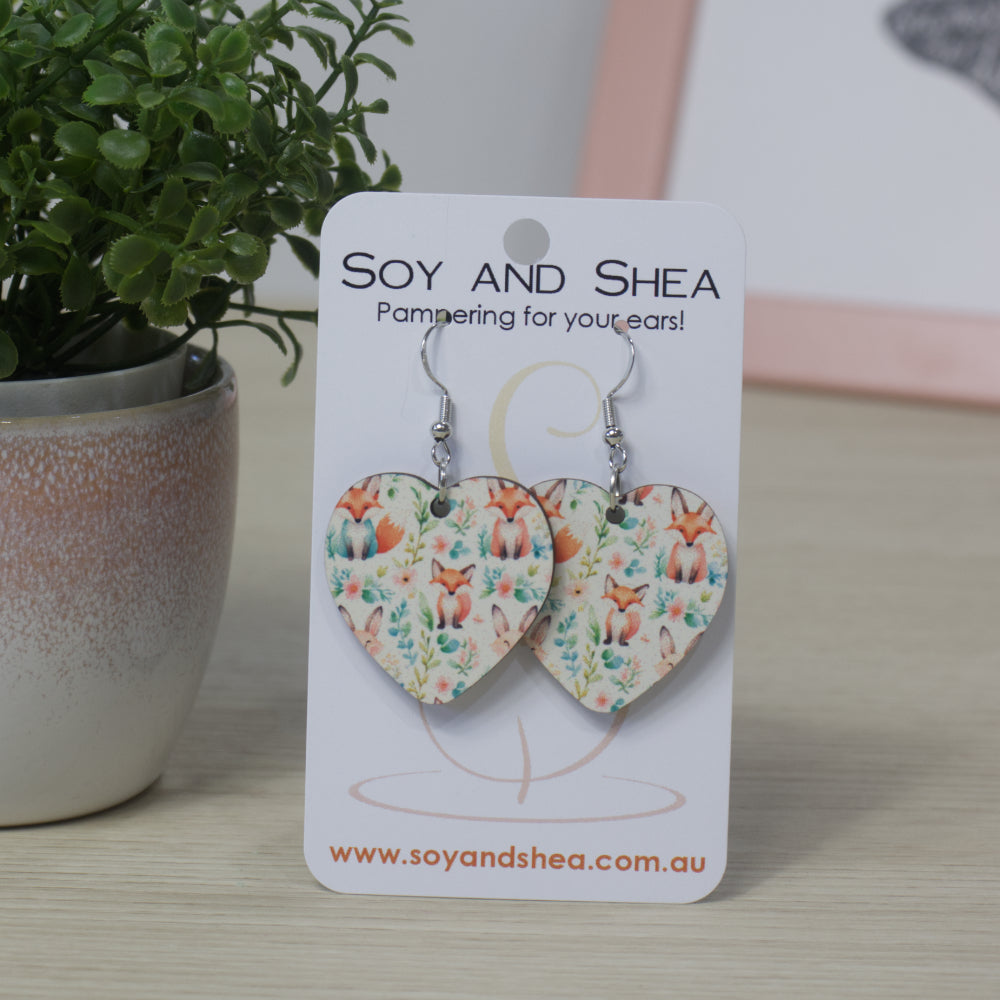 Heart-shaped earrings on a Soy and Shea card with a plant in the background.  The earrings feature a sublimated design of foxes sitting surround by flowers