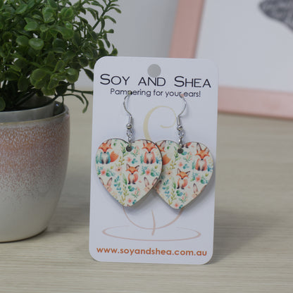 Heart-shaped earrings on a Soy and Shea card with a plant in the background.  The earrings feature a sublimated design of foxes sitting surround by flowers