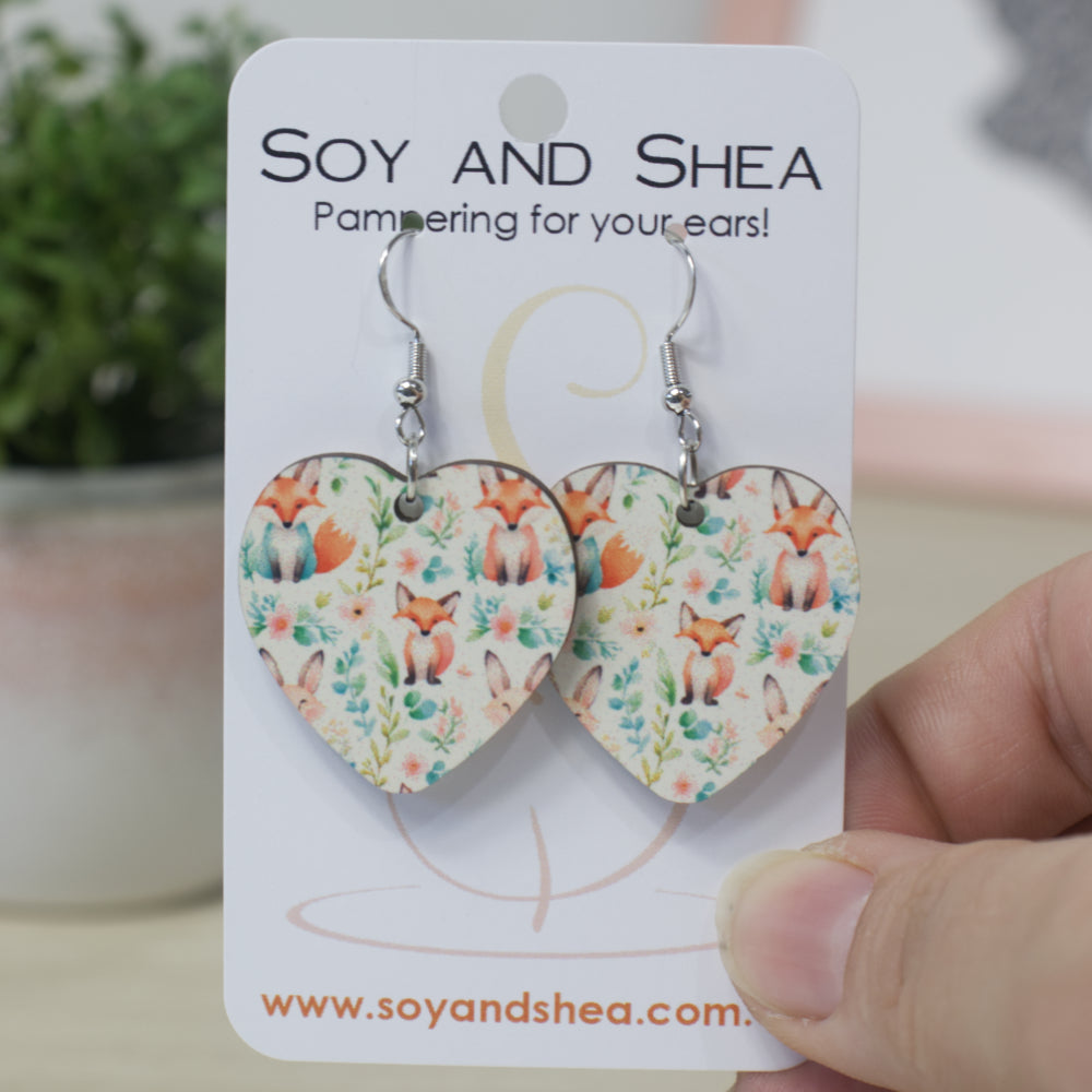 Heart-shaped earrings with silver hooks featuring a design of sitting foxes among flowers on a 'Soy and Shea' branded card.