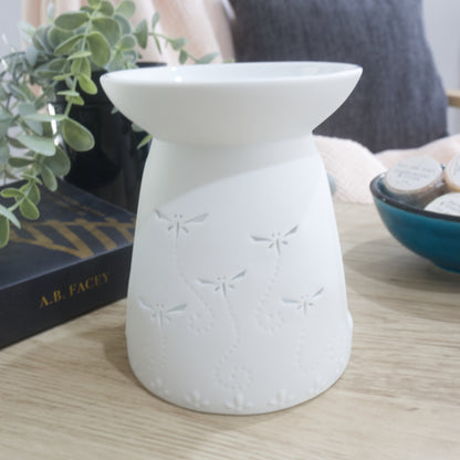 A matte white porcelain fragrance warmer sits on a light wooden table next to a blue bowl of wax melts and a box of matches. The warmer features embossed decorative details at the base and dragonfly cutouts. In the blurred background, a pink textured throw blanket and a dark grey cushion rest on a chair