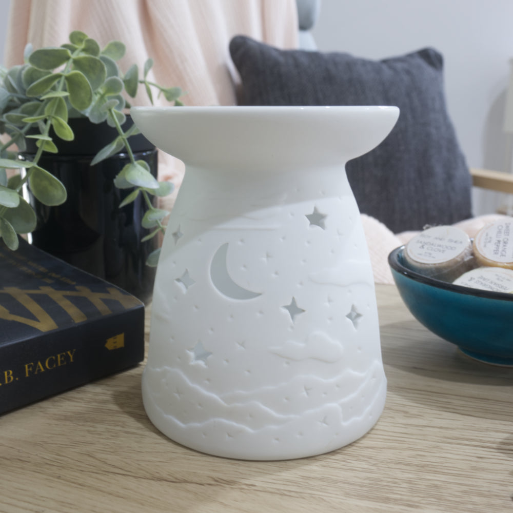 A matte white porcelain fragrance warmer sits on a light wooden table next to a blue bowl of wax melts and a box of matches. The warmer features embossed cloud details at the base with moon and stars cutouts. In the blurred background, a pink textured throw blanket and a dark grey cushion rest on a chair