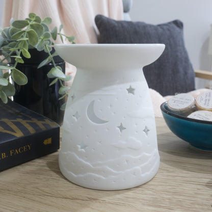 A matte white porcelain fragrance warmer sits on a light wooden table next to a blue bowl of wax melts and a box of matches. The warmer features embossed cloud details at the base with moon and stars cutouts. In the blurred background, a pink textured throw blanket and a dark grey cushion rest on a chair