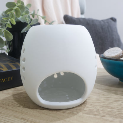 A smooth, unlit matte white ceramic fragrance warmer sits on a light wooden table, positioned to show its large, empty arch-shaped opening for a tealight candle. Part of a blue ceramic bowl filled with wax melts is visible on the right, and a black book and potted green foliage rest on the left. In the softly blurred background, a pink throw blanket and a dark grey cushion are draped over a chair.