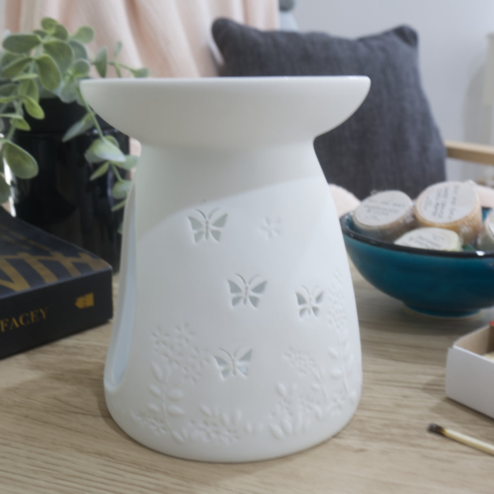 A matte white porcelain fragrance warmer sits on a light wooden table next to a blue bowl of wax melts and a box of matches. The warmer features embossed botanical details at the base and butterfly cutouts. In the blurred background, a pink textured throw blanket and a dark grey cushion rest on a chair