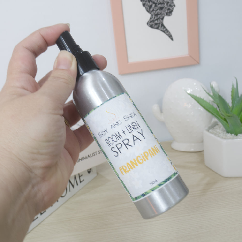 A hand holds a silver bottle with black spray cap over a bench with decorations like a plant and ornament.  The bottle is labeled and Room + Linen Spray Frangipani