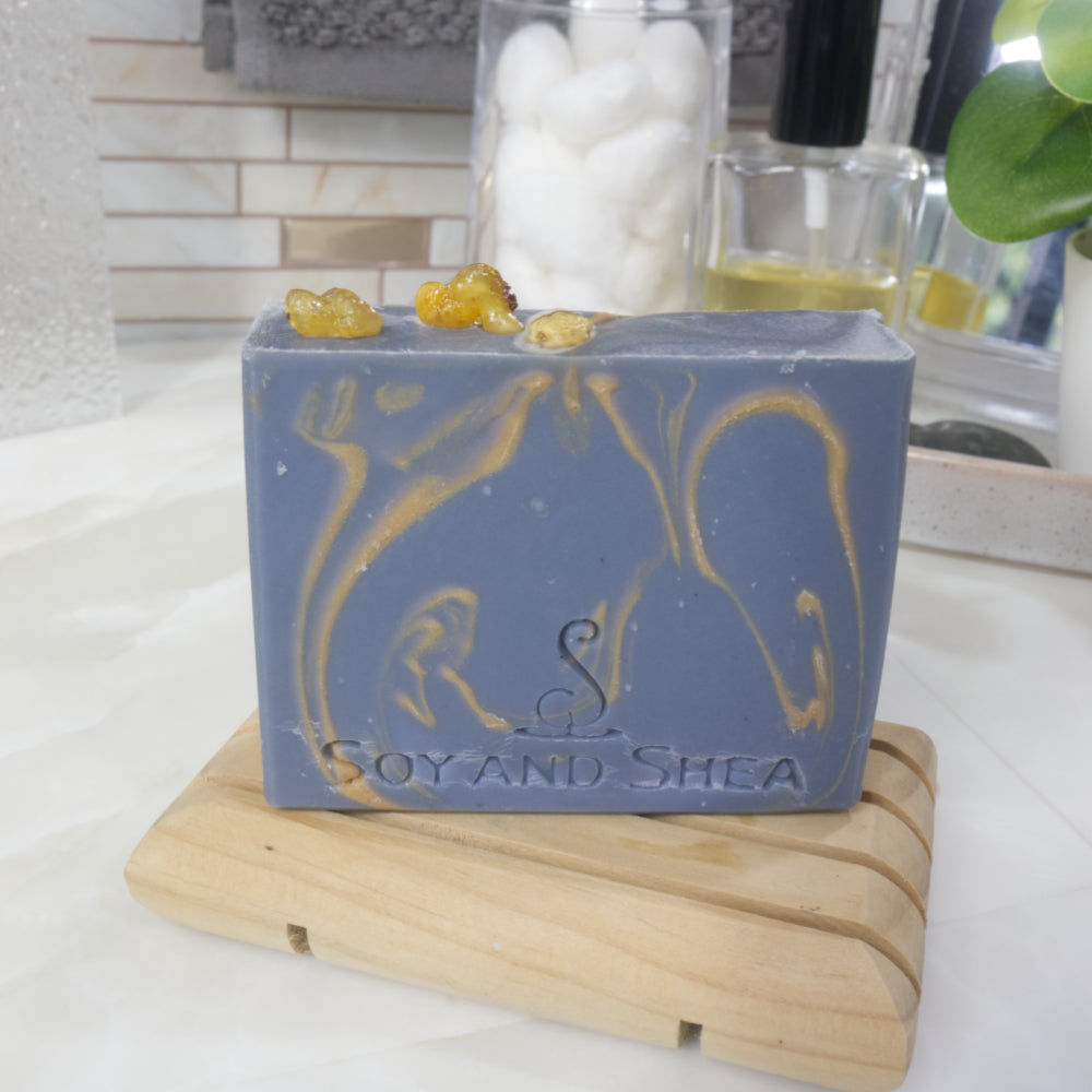 A dark blue soap with gold swirls stands upright on a wooden soap rack on a bathroom countertop.  In the background is a perfume bottle, a jar of cotton balls and greenery.   The top of the soap has three small pieces of resinous frankincense.