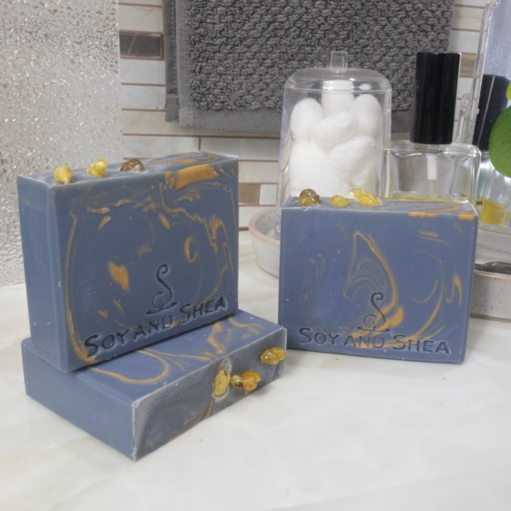 Three rectangular bars of soap are displayed on a bathroom countertop.  One soap lays on it side showcasing the top while another stands upright on it. A third soap stands upright to the right.  All soaps have a dark blue base colour with delicate gold swirls of colour.  Each soap has three small pieces of frankincense resins on the top.