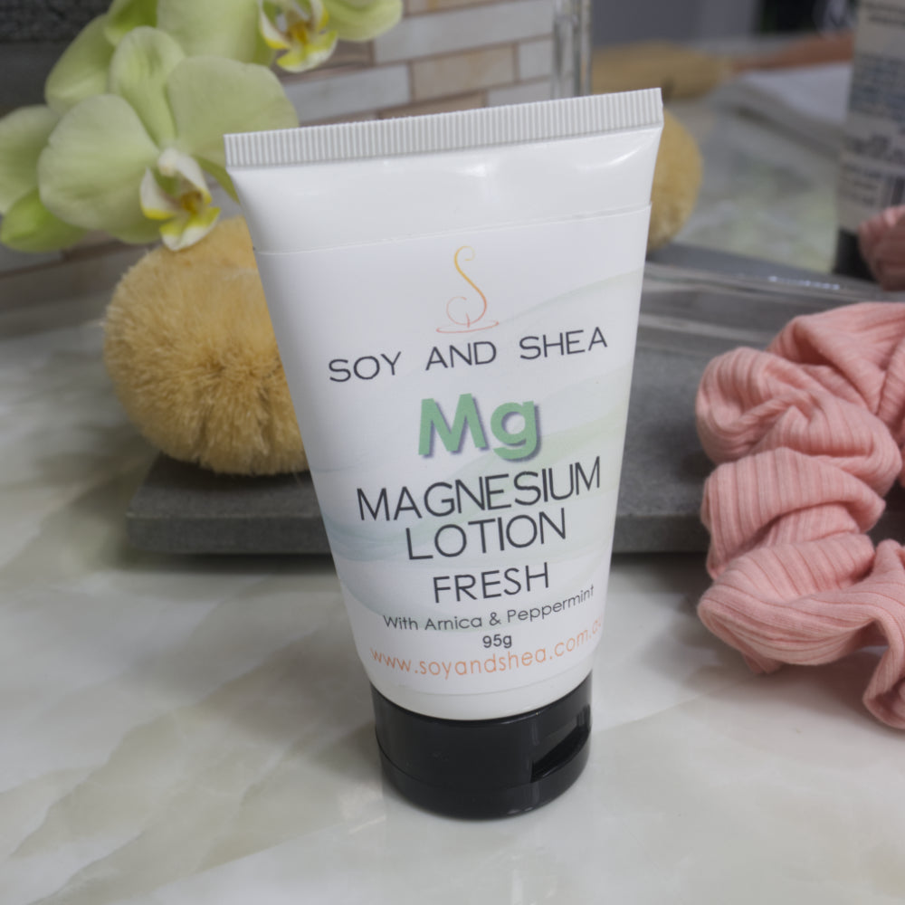 A white tube with black lid stands on a marble tile surface in front of a stone tray holding a mirror, body brush and scrunchie.   The tube has a white label with a faded green wavy pattern with the letters MG in green and under that MAGNESIUM LOTION FRESH in black capital letters.