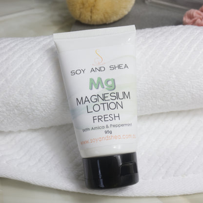 A white tube with black lid of Soy and Shea Magnesium Lotion Fresh  rests against a rolled up white towel with a bath sponge in the background.