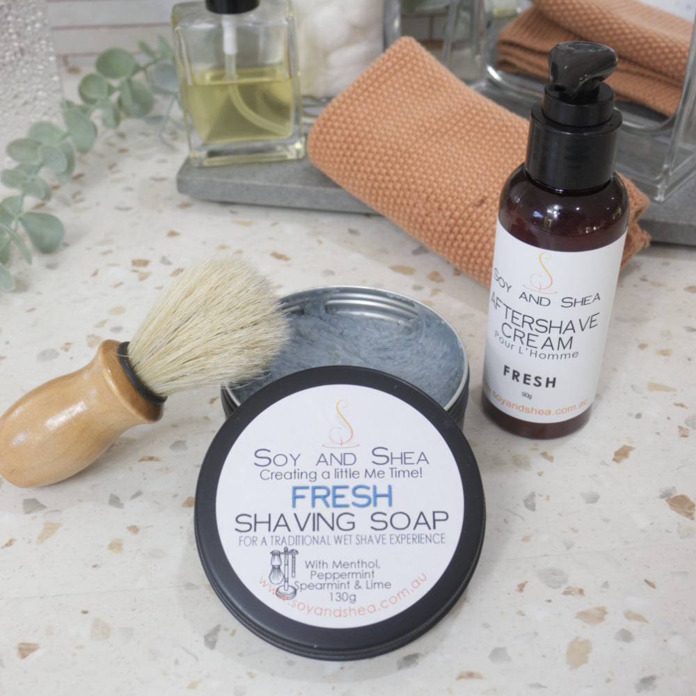 A black, round tin of shaving soap sits on a bathroom counter top with the lid off and resting on the side revealing a blue soap inside.  Resting against the side is shaving brush with wooden handle. On the other side of the tin is an amber bottle of Aftershave cream with lotion pump lid.