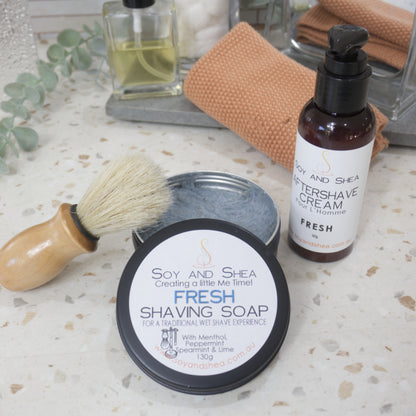 A black, round tin of shaving soap sits on a bathroom counter top with the lid off and resting on the side revealing a blue soap inside.  Resting against the side is shaving brush with wooden handle. On the other side of the tin is an amber bottle of Aftershave cream with lotion pump lid.