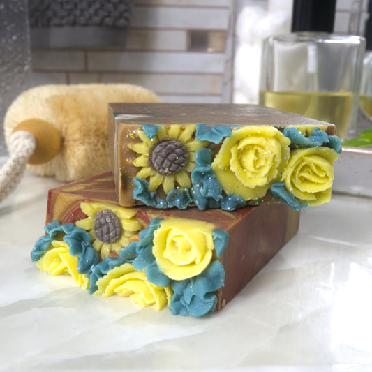 Two bars of soap lay flat on the bathroom countertop showing the decorative tops of the soap.  Each soap features a mix of sunflowers and yellow roses surrounded by green leaves.  These are all made of soap
