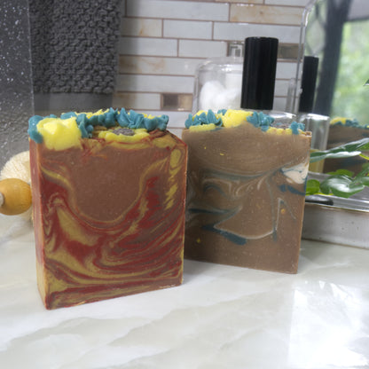 Two bars of soap sit on bathroom counter top.  The soap to the front left has swirls of orange, red and yellow, while the other to the right back has green and white swirls through a golden base.   The top has piped yellow flowers and green leaves