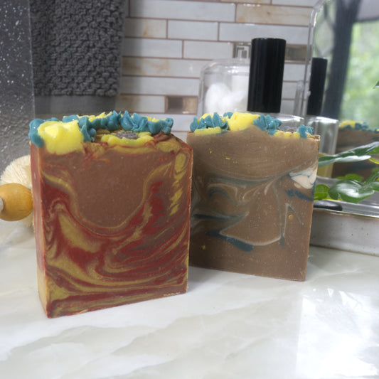 Two bars of soap sit on bathroom counter top.  The soap to the front left has swirls of orange, red and yellow, while the other to the right back has green and white swirls through a golden base.   The top has piped yellow flowers and green leaves