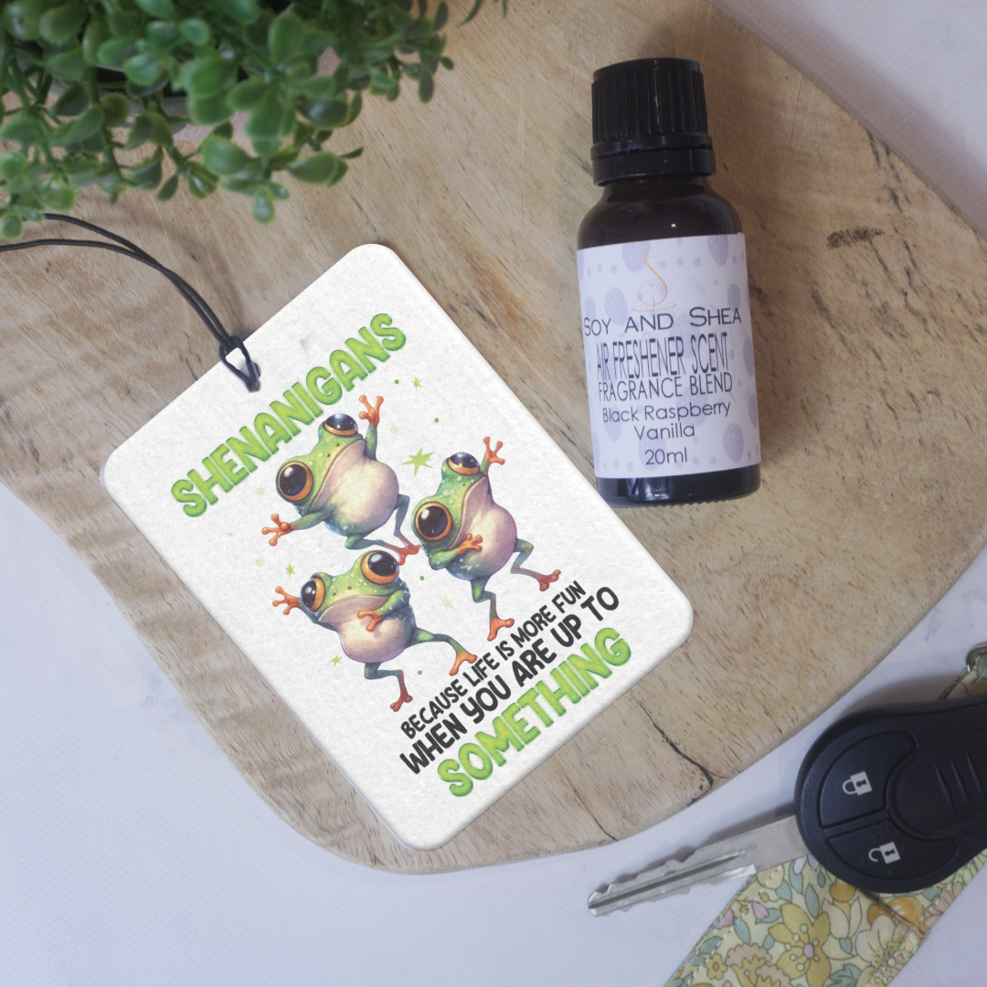 A car air freshener with a white background featuring a colorful print of three frogs and the text 'SHENANIGANS because life is more fun WHEN YOU ARE UP TO SOMETHING' lies on a wooden try next to bottle of Air Fresh Scent