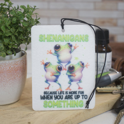 A car air freshener with a white background featuring a colorful print of three frogs and the text 'SHENANIGANS because life is more fun WHEN YOU ARE UP TO SOMETHING'.  The air freshener leans agains a potted plant and has a set of keys next to it.