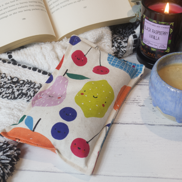 A mini rectangular heat pack rests against a black and white blanket and open book.  Next to it is a burning candle and filled coffee cup evoking a comfy scene.    The heat pack has a cream coloured fabric decorated with drawings of various fruit including cherries, blueberries, pear and lemon all having a small smiley face.