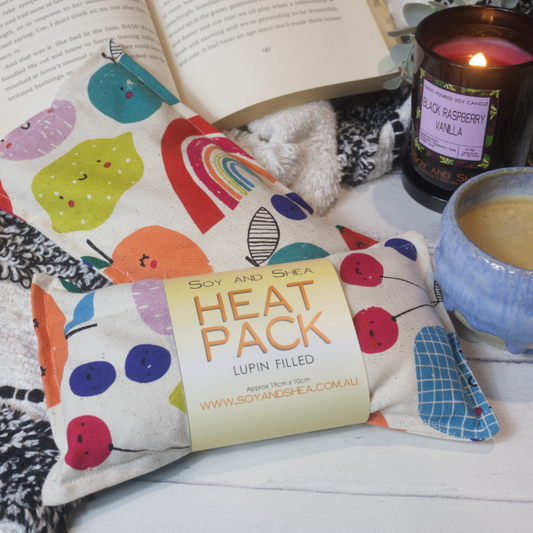 Two Mini rectangular heat packs are stacked on each other, one with a simple belly band label reading HEAT PACK LUPIN FILLED.  They have a cream fabric and each show a different design featuring basic illustrations of various fruits with small smiley faces.  The fruits include lemons, orange, cherries and blueberries.   The heat packs rest against a black and white blanket and open book.  Next to it is a burning candle and filled coffee cup evoking a comfy scene.  