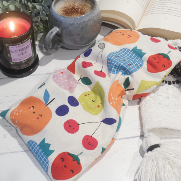 A small rectanglular Heat Pack rests on a wooden table top along with a black and white blanket, open book, burning candle and filled coffee cup evoking a cosy scene.  The Heat pack has a cream fabric decorated with simple drawings of various fruits including cherries, orange, pineapple and blueberries. All fruit feature a smiling face.
