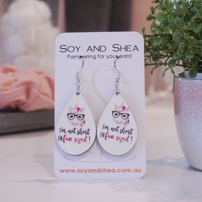 A pair of earrings on silver hooks hang on a white card branded with SOY AND SHEA leaning against a pot with a green leafy plant and a blurred background.  The earrings are teardrop shaped with an illustration of a unicorn with a rainbow mane and glasses towards the top of the teardrop with the text I'M NOT SHORT I'M FUN SIZED underneath