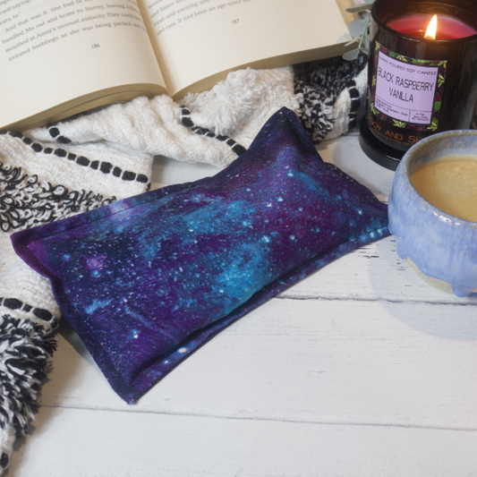 A mini rectangular heat pack rests against a black and white blanket and open book.  Next to it is a burning candle and filled coffee cup evoking a comfy scene.   The heat pack has a marbled galaxy sky in shades of blue and purple with dots of white to represent stars