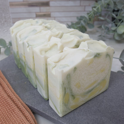 A row of soap bars sit on a grey stone tray with a tiled background and greenery.  The front bar of soap is white with splashes of dark green and yellow throughout and features a textured top