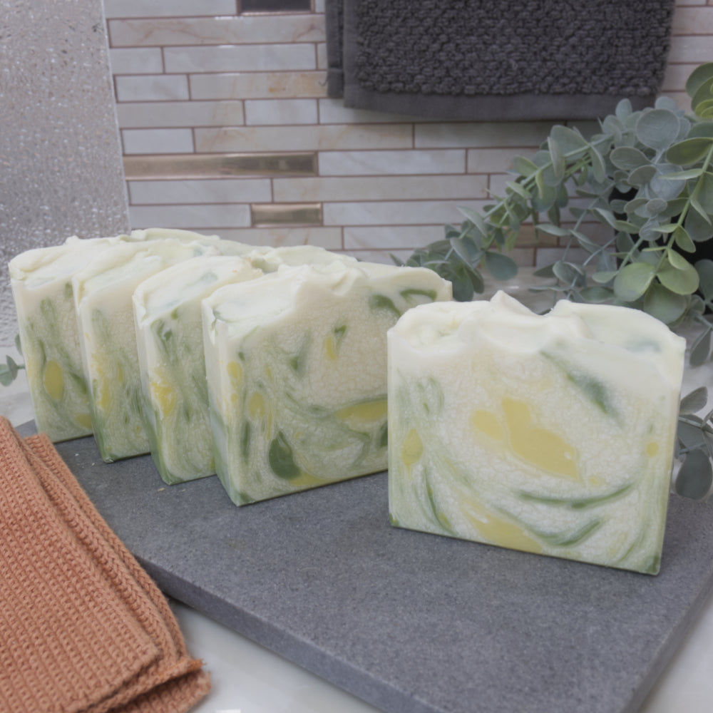 A selection of five soaps sit in a row one in front of the other on a grey stone tray.  The soaps are offset slightly to show a peak of the design on each soap.  The soap at the front shows they are white with splashes of green and yellow.  These are surrounded by greenery and towels