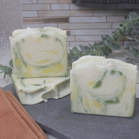 Three bars of soap sit on a gray stone tray with a tiled wall and plant in the background.  One soap lays flat on the stone surface showing the textured wavy top.  Another stands on top of this soap showing a whote soap with drops of green and yellow drops. In front is another soap with the same colours but different pattern