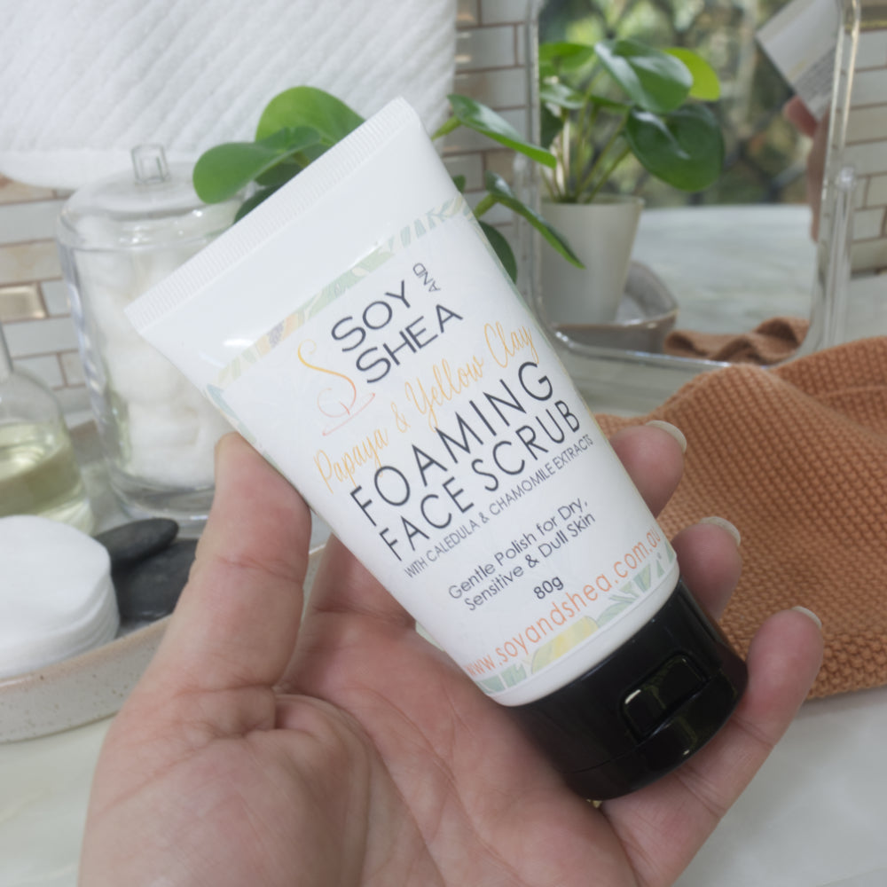 A hand holding a white squeeze tube of Soy & Shea Papaya & Yellow Clay Foaming Face Scrub, with a bathroom countertop and plants blurred in the background.