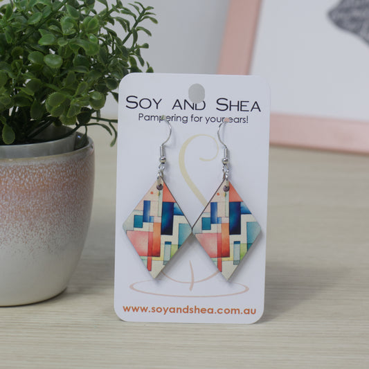 Diamond shaped earrings with a colourful geometric design hang on a Soy and Shea card with a plant in the background