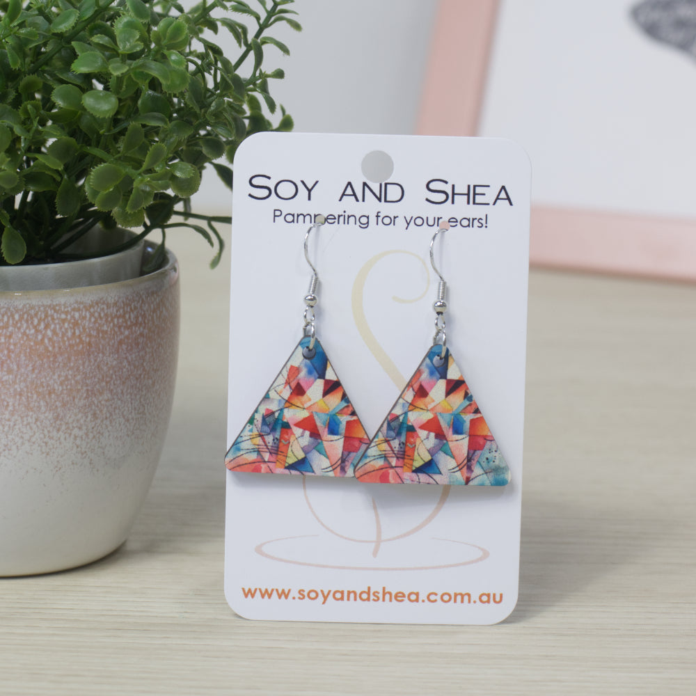 Triangle shaped earrings with a colourful geometric design on a Soy and Shea display card with a plant in the background.