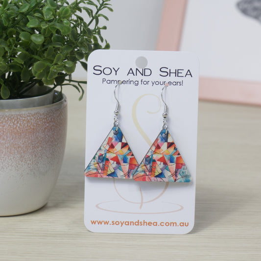 Triangle shaped earrings with a colourful geometric design on a Soy and Shea display card with a plant in the background.