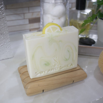 A bar of soap stands on a wooden soap rack on a bathroom countertop.  The soap is white with fine swirls of yellow and green throughout.  On the top of the soap is a lemon slice standing upright in the center.  It is made with soap