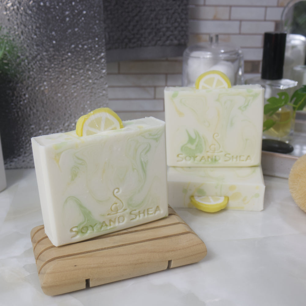 Three soap bars are displayed on a bathroom counter top.  One soap lays flat show casing the top of the soap with another soap standing on top of it. To the front left is a third soap standing on a wooden soap rack.  All the soaps are white with varying swirls of green and yellow throughout. Each soap features a lemon slice made of soap sticking into the top center of the soap bar