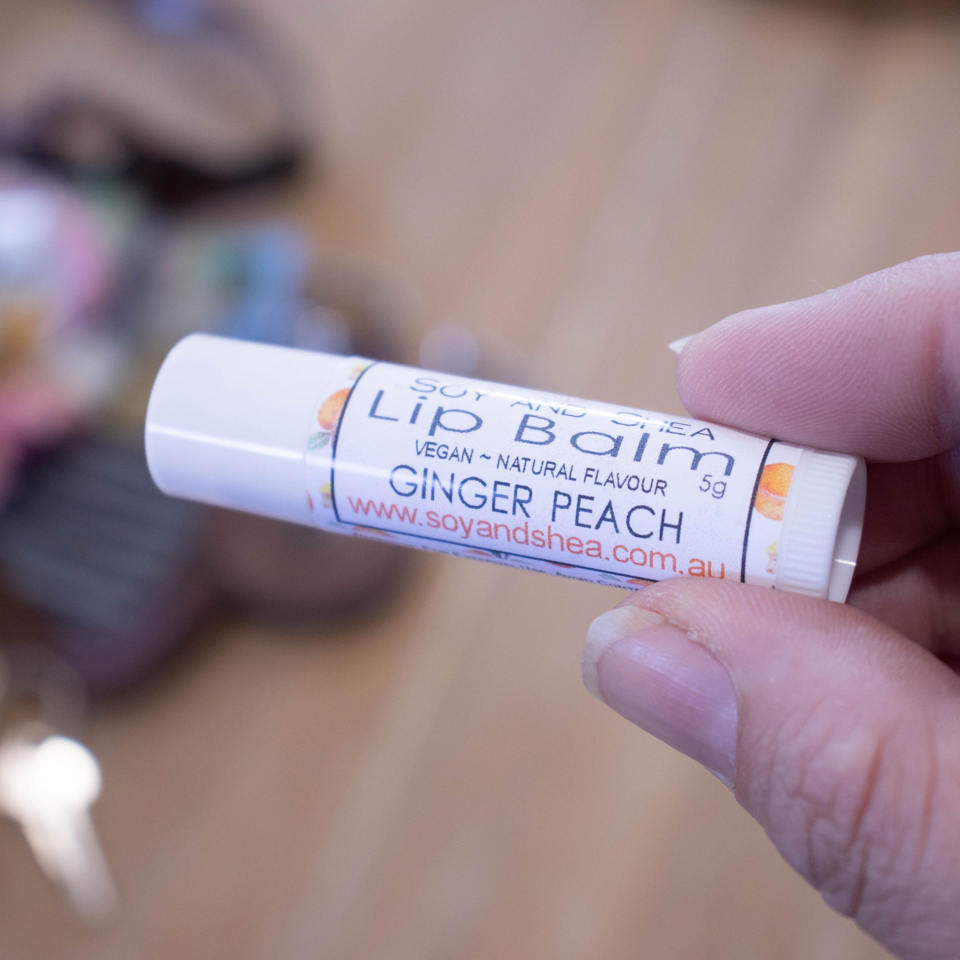 A hand holds a white tube labelled lip balm over a blurred image of an open handbag with contents spilling onto the table.  The lip balms features a white label with watercolour images of peaches and ginger slices