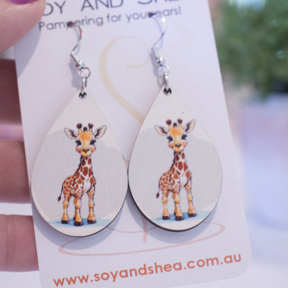 A hand holds a white card branded with SOY AND SHEA with a pair of earrings hanging on hooks.  The earrings are teardrop shaped with an illustration of a giraffe standign against a watercolour grey background