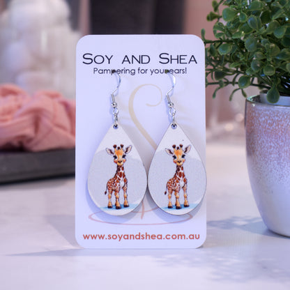 A pair of earrings on silver hooks hang on a white card branded with SOY AND SHEA leaning against a pot with a green leafy plant and a blurred background.  The earrings are teardrop shaped with a girraffe standing against a grey and white background