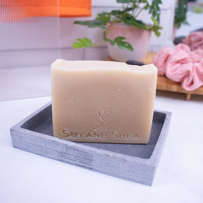 A beige colored soap bar labeled 'Soy and Shea' placed on a gray dish with a blurred background featuring plants and a pink cloth.