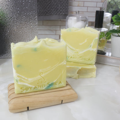 Three soap bars are displayed on a bathroom counter top.  One soap lays flat show casing the top of the soap with another soap standing on top of it. To the front left is a third soap standing on a wooden soap rack.  All the soaps have a bright yellow base colour with streaks of white and green throughout.  The stop of the top has been finished with a wavy top.