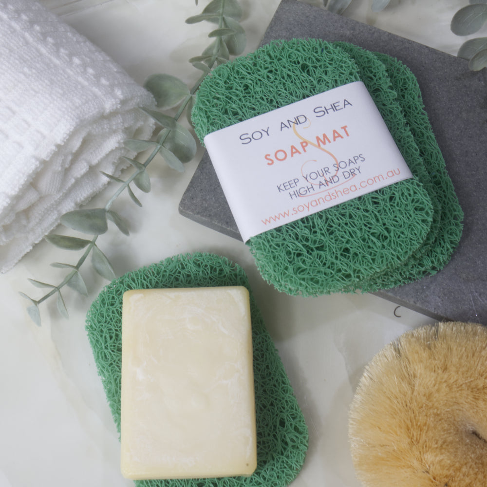 A flatlay image showing a collection of Green Soap mats, displayed on a bathroom countertop along with a white towel, body brush and greenery.  Three soap mats are stacked upon each other in a fan shape on a grey stone tray with the top one featuring a product label.  A fourth mat lies to the bottom of the image with a cream soap lying on it.