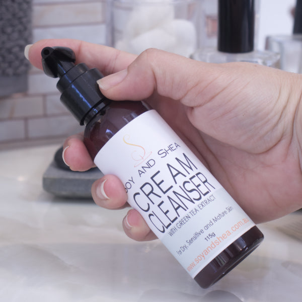 A hand holds a bottle with pump lid of Soy and Shea Cream Cleanser, formulated for sensitive and mature skin, in a bathroom with tiled walls and assorted toiletries.