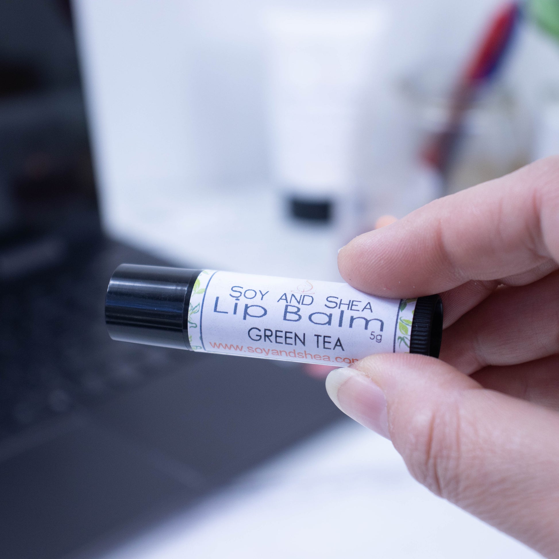 A hand holds a black tube of lip balm against a blurred background with an open laptop computer on a white surface.  The label has text LIP BALM GREEN TEA on a white background with a border showing green leaves