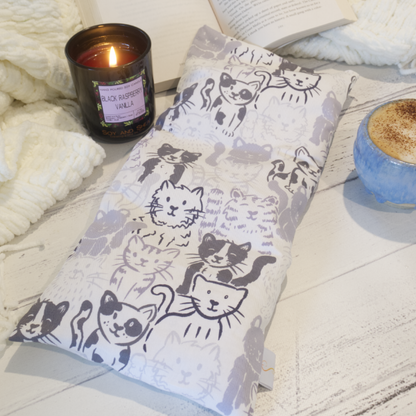 A heat pack featuring pencil drawinging of grey cats sitting side by side and in rows is placed on a wooden surface resting agains an open book and white blanket, accompanied by a burning candle and a cup of coffee.  