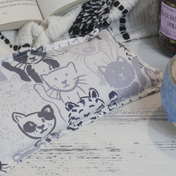A close up shot of the Grey Cats Mini Heat pack showing a variety of cats sketched in tones of grey on a white background.  The pack rests on wooden surface surround by a blanket and open book