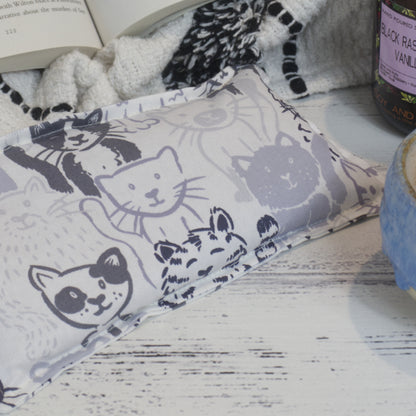 A close up shot of the Grey Cats Mini Heat pack showing a variety of cats sketched in tones of grey on a white background.  The pack rests on wooden surface surround by a blanket and open book