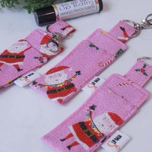 A selection of three clip balms showing variations in fabric design, lie on a wooden side with the middle Clip Balm attached to a car key.  A lip balm lays near-by along with some greenery.  The Clip balms are bright pink with flecks of gold and feature drawings of santa and candy canes.