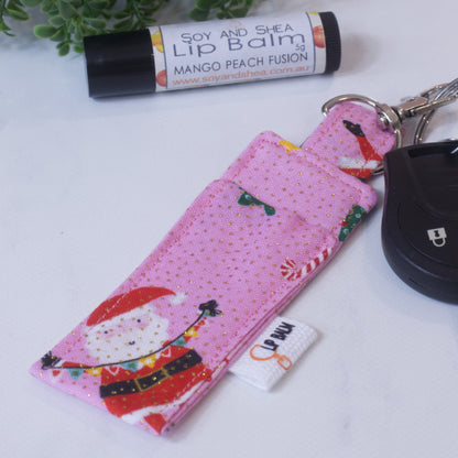 A lip balm holder lies on a wooden surface attached to a car key with a "lobster claw" clip.  To the side is a tube of lip balm surrounded by greenery.  The Clip Balm is bright pink with gold dots and features a cartoon image of a santa and candy cane